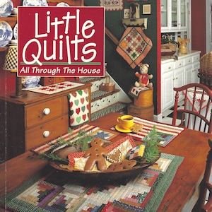 May include: A book cover titled Little Quilts All Through The House by Alice Berg, Mary Ellen Von Holt, and Sylvia Johnson. The cover features a wooden table with a patchwork runner, a bowl of gingerbread cookies, and a yellow cup and saucer.