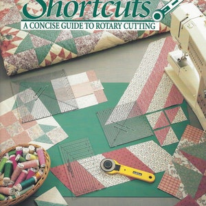 Op de afbeelding: Een boek getiteld "Shortcuts: A Concise Guide to Rotary Cutting" door Donna Lynn Thomas. De omslag toont een patchworkdeken, roterende snijgereedschappen en stofresten.