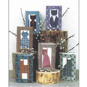 May include: A collection of six framed quilted cat designs in various colors and patterns, including plaid, gingham, and floral prints. The frames are made of wood and have a rustic finish. The title "Kitty Cover-Ups" and the text "the Plaid Cat" are visible below the frames.