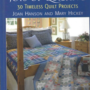 May include: A book titled The Simple Joys of Quilting: 30 Timeless Quilt Projects by Joan Hanson and Mary Hickey. The cover features a photograph of a wooden four-poster bed covered in a colorful patchwork quilt with blue floral pillows.