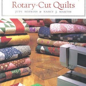Puede incluir: Un libro titulado 101 Fabulous Rotary-Cut Quilts de Judy Hopkins y Nancy J. Martin, con pilas de colchas estampadas de colores, una máquina de coser gris claro, dos reglas acrílicas transparentes y un cortador rotativo amarillo sobre una base.