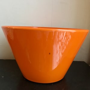 May include: A large, bright orange ceramic bowl with a wide, rounded base and a flared lip. The bowl is glazed and has a smooth, glossy finish.