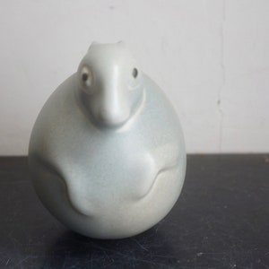 May include: A ceramic sculpture of a stylized animal, possibly a rabbit or a bear, with a rounded body and a small head. The sculpture is painted in a light gray color and has a smooth, matte finish.