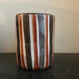 May include: A cylindrical ceramic vase with vertical stripes in alternating colours of dark brown, white, and reddish-brown. The vase has a slightly textured surface and a simple, modern design. The vase is suitable for holding flowers or as a decorative item.
