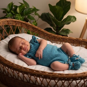 Könnte beinhalten: Ein schlafendes Baby in einer Korbbassinet. Das Baby trägt einen gestrickten türkisfarbenen Strampler mit passenden Babyschuhen. Die Bassinet ist mit einem weißen Laken ausgekleidet, und der Hintergrund zeigt grüne Pflanzen und eine warme Lampe.