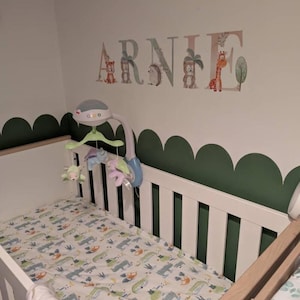 May include: A white wooden cot with a patterned sheet featuring small animal illustrations. Above the cot, a wall decal displays the name ARNIE in large letters decorated with jungle animals and trees. A mobile with hanging plush toys is attached.