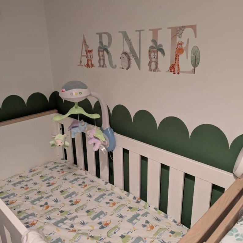 May include: A white wooden cot with a patterned sheet featuring small animal illustrations. Above the cot, a wall decal displays the name ARNIE in large letters decorated with jungle animals and trees. A mobile with hanging plush toys is attached.