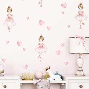 May include: A light pink and white wall decal featuring illustrations of ballerinas and pink hearts. The ballerinas are wearing pink dresses and ballet shoes. The background is a light pink colour, creating a whimsical and charming aesthetic.