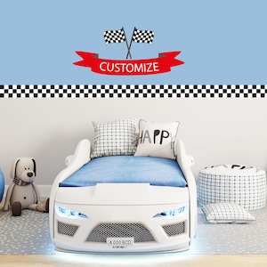 May include: A white race car bed with blue bedding, a checkered flag banner, and a pillow that says "HAPPY." The bed has a front grill, headlights, and a license plate. A stuffed dog and a patterned ottoman are also in the room.