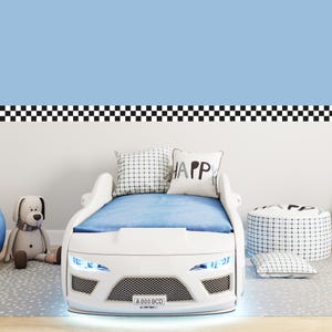 Op de afbeelding: Een wit autovormig bed met een blauwe matras en kussens. Het bed heeft koplampen en een kentekenplaat met "A 000 BCD". Een knuffelhond en een poef met patroon staan ook in de kamer. De muur is lichtblauw met een zwart-wit geblokte rand.