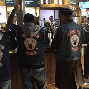 May include: Four people wearing denim jackets with black spider patches that say "Black Widows Paguma" are standing in a pub. They are all wearing jeans and have a casual look.