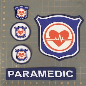 May include: Four blue and white embroidered patches with a red heart and a heartbeat line inside a circle. The largest patch is a shield shape. The smaller patches are round. A blue and white fabric strip with the word "PARAMEDIC" in white letters.