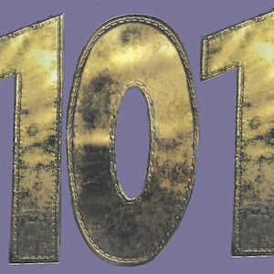 Puede incluir: Un número "101" de color dorado con una textura y una costura visibles. Los números están delineados en un color dorado más oscuro.