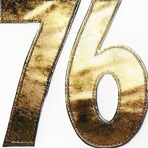 Puede incluir: Número 76 metálico dorado con un contorno cosido. Los números son grandes y tienen una textura ligeramente en relieve.