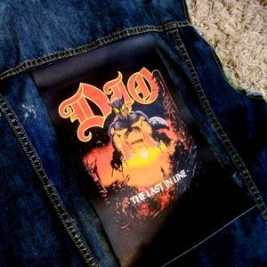 May include: A dark blue denim waistcoat featuring a large rectangular patch on the back. The patch displays the Dio logo in orange and yellow, with a central horned figure, a glowing orange horizon, and the text THE LAST IN LINE printed below.