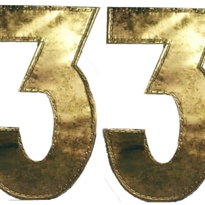Könnte beinhalten: Zwei goldfarbene, gestickte Stoffzahlen, jede eine "3", bilden die Zahl "33".