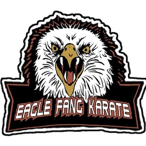 May include: A black and white illustration of an eagle's head with a fierce expression. The eagle's beak is open and its eyes are wide. The eagle's head is surrounded by a brown and white border. Below the eagle's head is a black banner with the text "Eagle Fang Karate" in white letters.