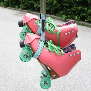 Könnte beinhalten: Ein Paar rosa Rollschuhe mit grünen Rollen und Schnürsenkeln. Die Schuhe sind an einem grün-weißen Band befestigt, auf dem der Text "Voodoocollections" steht.