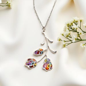 Puede incluir: Un collar plateado con un colgante floral. El colgante tiene tres elementos en forma de flor con un efecto vidriera, con una mezcla de colores rojo, morado, amarillo y azul. El collar se muestra sobre un fondo blanco.