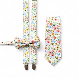 Puede incluir: Un conjunto de accesorios florales que incluye tirantes, pajarita y corbata. La tela blanca presenta un estampado de flores rosas, amarillas y azules con hojas verdes. Los tirantes tienen clips marrones. La pajarita está preatada.