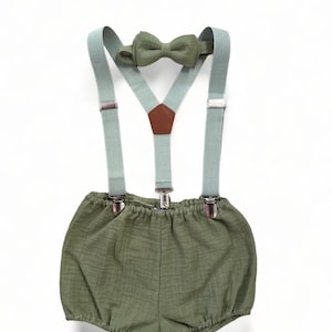 Może przedstawiać: Zestaw zielonych dziecięcych szelków z dopasowaną muszką. Szelki mają srebrne klipsy i brązowy skórzany detal. Muszka jest wykonana z miękkiej tkaniny.