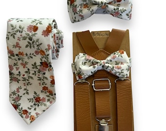 Puede incluir: Un conjunto de tirantes de cuero marrón con una pajarita floral y una corbata a juego. La pajarita y la corbata tienen un fondo blanco con flores naranjas y rosas.