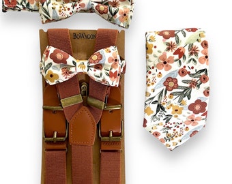 Puede incluir: Un conjunto de tirantes marrones con una pajarita floral y una corbata a juego. La pajarita y la corbata tienen un fondo blanco con flores rosas, naranjas y amarillas. Los tirantes tienen detalles de cuero y hebillas doradas. El conjunto está empaquetado en una tarjeta marrón con la palabra "BOWAGON" impresa en ella.