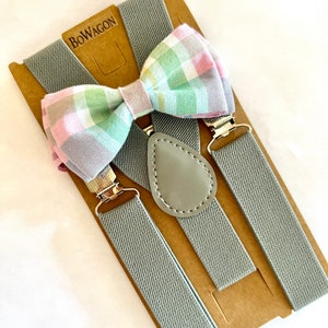 May include: Gray suspenders with a bow tie. The bow tie is a plaid pattern with pink, green, and white. The suspenders have a leather detail and the brand name "BOWAGON" is printed on the packaging.