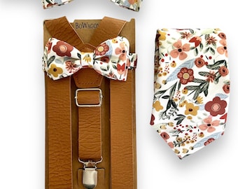 Puede incluir: Un conjunto de tirantes de cuero marrón con una pajarita floral y una corbata floral a juego. La pajarita y la corbata tienen un fondo blanco con flores rojas, rosas y amarillas.