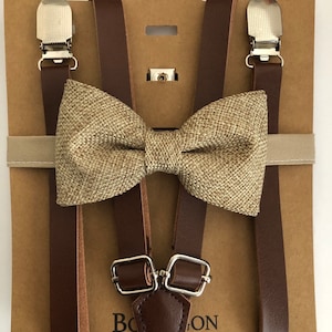 May include: A brown leather suspender set with a matching bow tie. The bow tie is made of a textured tan fabric. The suspenders have silver-coloured metal clips and adjustable buckles. The set is displayed on a brown cardboard backing.