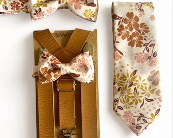 Puede incluir: Un conjunto de tirantes de cuero marrón con una pajarita floral y una corbata a juego. La pajarita y la corbata tienen un fondo blanco con patrones florales rosas y marrones. Los tirantes están empaquetados en una caja de cartón marrón con el nombre de la marca "BoWagon" impreso en ella.