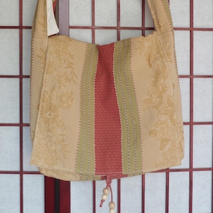 May include: A beige shoulder bag with a floral pattern and a red and green striped center panel. The bag has a long strap and a drawstring closure with wooden beads. The background is a red and white grid.