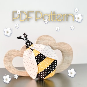 May include: A PDF pattern for sewing a cute bee toy. The bee is yellow and black with white polka dots and has a pink and white wing. The bee is sitting on a wooden heart-shaped base.