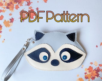 Puede incluir: Un bolso de color azul claro con forma de mapache y una correa negra. El bolso tiene detalles en fieltro blanco y negro y botones de ojos azules. El texto "PDF Pattern" está escrito en letras naranja y amarillas en la parte superior de la imagen.