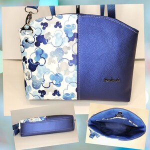 May include: A blue and white tote bag with a Mickey Mouse print on one side and a solid blue side. The bag has a top zipper closure and a detachable shoulder strap. The bag is handmade and has the word "remarkable" embossed on the blue side.