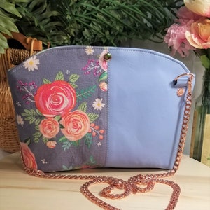 May include: A light blue crossbody bag with a floral print on one side and a solid color on the other. The bag has a rose gold chain strap.