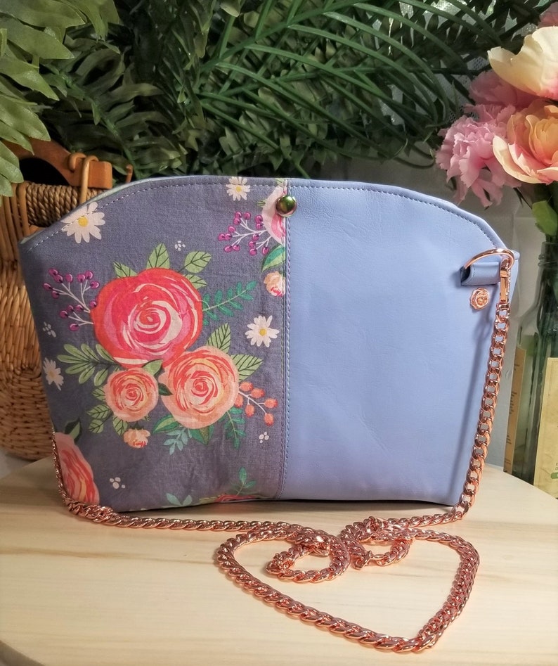 May include: A light blue crossbody bag with a floral print on one side and a solid color on the other. The bag has a rose gold chain strap.