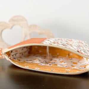 May include: A small, zippered pouch with a floral print on one side and a solid orange fabric on the other. The pouch is open and the inside is lined with a yellow floral fabric.