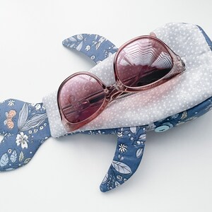 Puede incluir: Una funda de tela azul en forma de ballena con un patr&oacute;n floral y un cierre de bot&oacute;n. La funda contiene un par de gafas de sol rosas.