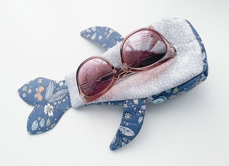 Puede incluir: Una funda de tela azul en forma de ballena con un patr&oacute;n floral y un cierre de bot&oacute;n. La funda contiene un par de gafas de sol rosas.
