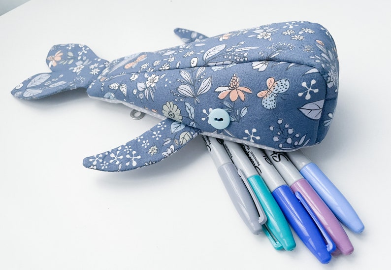Puede incluir: Un estuche para l&aacute;pices con forma de ballena azul con estampado floral y un bot&oacute;n azul claro. El estuche est&aacute; abierto y lleno de rotuladores de colores.