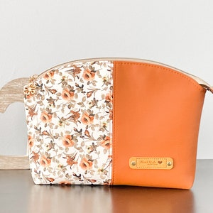 May include: A white and orange zippered pouch with a floral print on one side and a solid orange color on the other side. The pouch has a gold zipper and a gold tag that says "Hand Made".