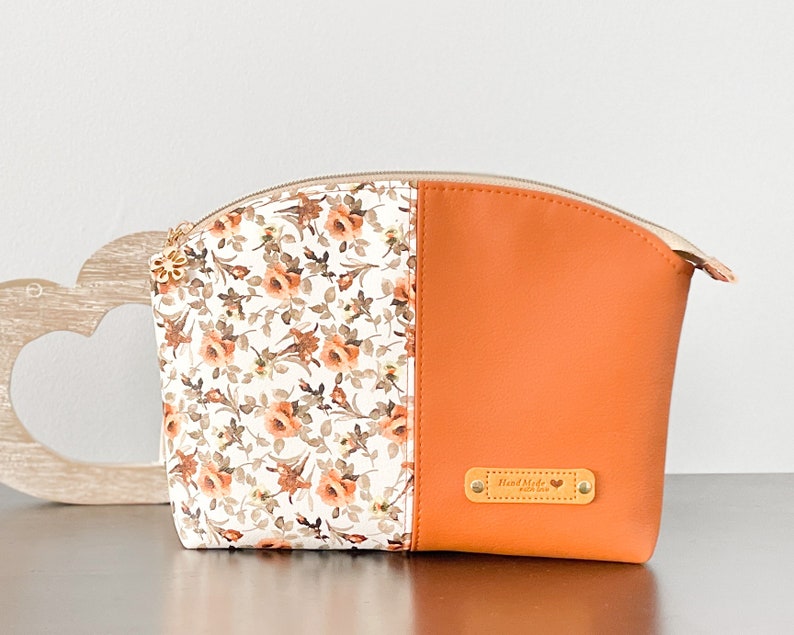 May include: A white and orange zippered pouch with a floral print on one side and a solid orange color on the other side. The pouch has a gold zipper and a gold tag that says "Hand Made".
