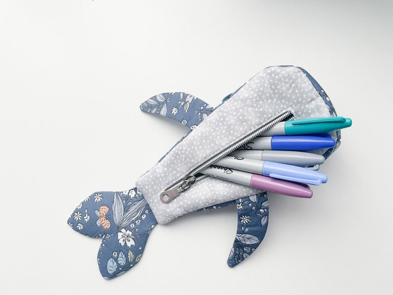 Puede incluir: Un estuche para l&aacute;pices con forma de ballena azul con estampado floral y cierre de cremallera. El estuche est&aacute; lleno de rotuladores de colores.