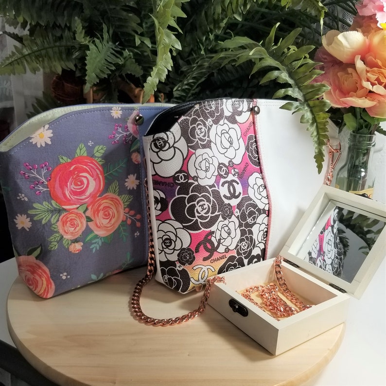 May include: Three tote bags with floral patterns. The bag on the left is blue with pink and white flowers. The bag in the middle is white with black and pink flowers and the text "CHANEL". The bag on the right is white with a floral pattern. A small white box with a mirror lid and a gold chain necklace are in the foreground.