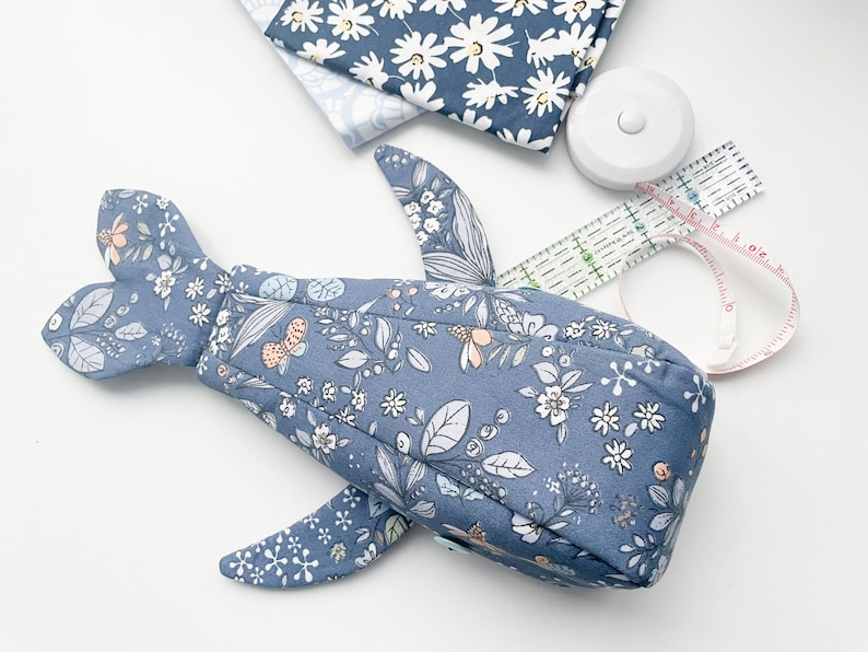 Puede incluir: Una ballena de tela azul con un patr&oacute;n floral. La ballena tiene un cuerpo largo y una aleta de cola. La tela es de color azul claro con un patr&oacute;n floral blanco.
