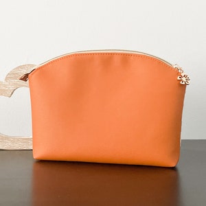 May include: A bright orange zippered pouch with a small gold flower charm. The pouch is made of a smooth, faux leather material.