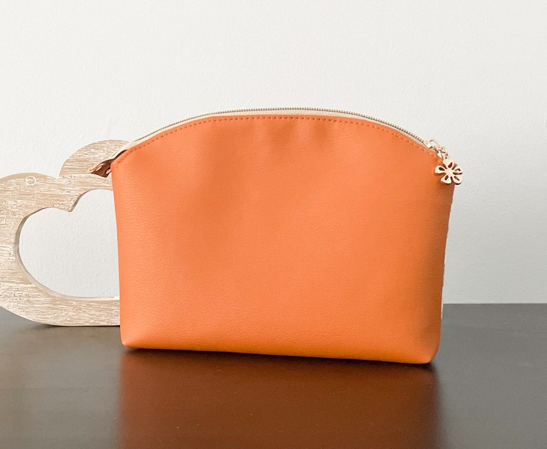 May include: A bright orange zippered pouch with a small gold flower charm. The pouch is made of a smooth, faux leather material.