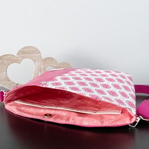 May include: A pink and white patterned crossbody bag with a pink strap. The bag is open and the inside is lined with a pink fabric.