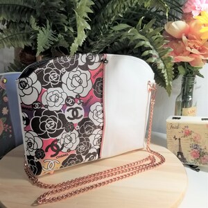 May include: A white and black floral print handbag with a rose pattern and the word "Chanel" repeated throughout. The bag has a white leather flap and a rose gold chain strap.